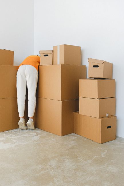 A person dressed in white trousers and an orange top is seen inside a room, standing next to a cluster of neatly packed cardboard moving boxes in various sizes, some with built-in handles. The boxes a