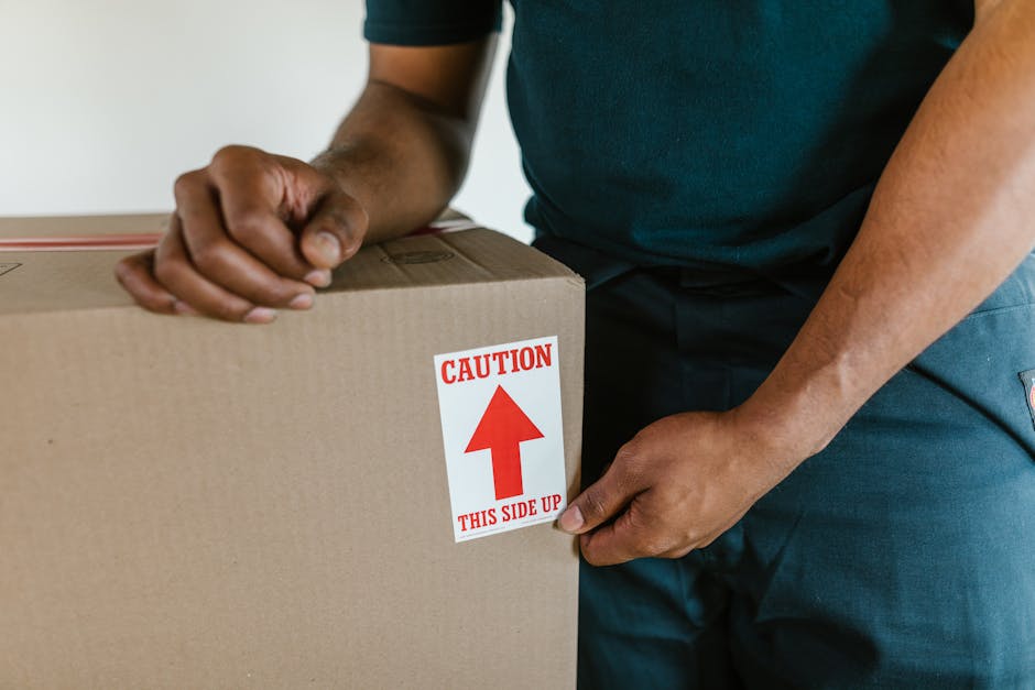 A person dressed in white trousers and an orange top is seen inside a room, standing next to a cluster of neatly packed cardboard moving boxes in various sizes, some with built-in handles. The boxes a