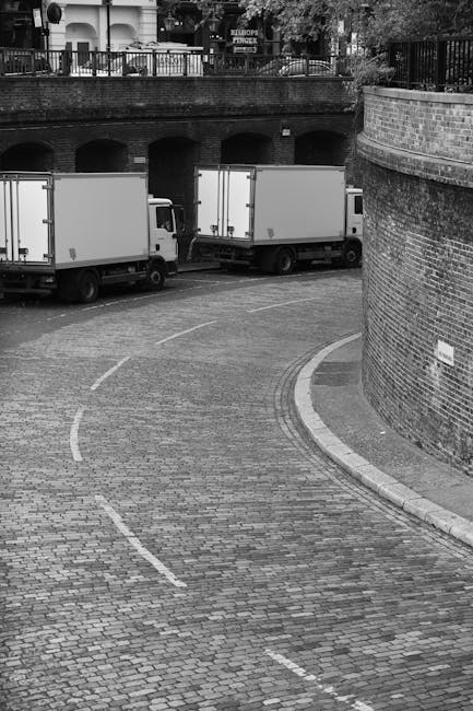 A black and white photograph shows two white moving trucks parked on a cobblestone driveway adjacent to a brick wall with a curved corner. The trucks are positioned side by side, facing the same direction, with their cargo areas visible. The driveway curves to the right, leading towards an underpass or tunnel beneath a bridge, which is supported by brick columns. Above the bridge is a metal railing and a row of trees and buildings can be seen in the background. The scene suggests a home relocation or furniture transport process, with the trucks ready for a house removal in Ickenham, as operated by Man With a Van Ickenham. The lighting appears natural, with no visible shadows, indicating an overcast day. The space is clear of pedestrians or other vehicles, emphasizing the loading or unloading area for household items and boxes prepared for transportation.