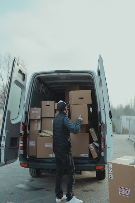 A man wearing a dark jacket, trousers, and a black cap is standing beside an open white moving van in an outdoor parking area, surrounded by scattered cardboard boxes of various sizes, some sealed with packing tape and labeled, which are stacked inside the van. The interior of the van shows a spacious cargo area used for furniture transport and home relocation purposes, with additional boxes visible in the background. The man appears to be in the process of loading or organizing the boxes, some of which are positioned on the ground nearby, including one marked as fragile. The scene is set during daytime with a cloudy sky, and the background includes a few industrial or storage units, indicating a relocation or delivery operation by a professional removals service, such as Man With a Van Ickenham, focusing on packing, moving, and home logistical support.