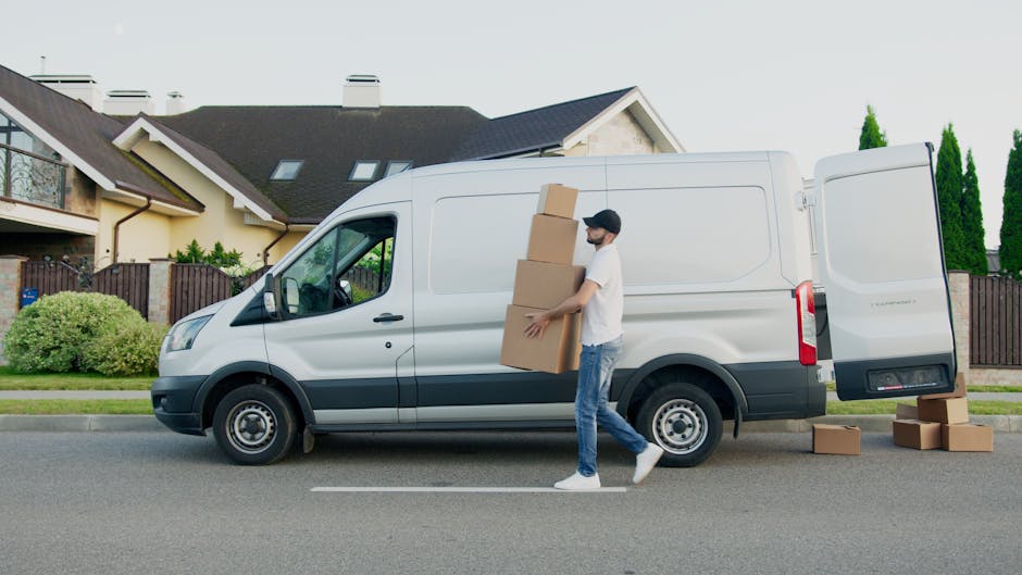 A man wearing a white shirt and jeans is standing beside an open white moving van parked on a residential street in Ickenham. He is in the process of packing or unpacking cardboard boxes of various si