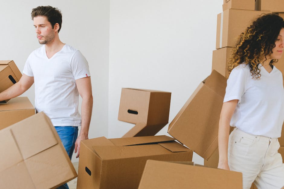 Two individuals involved in packing and home relocation activities indoors, surrounded by stacked cardboard moving boxes of various sizes made from plain brown material with black handles. The man on