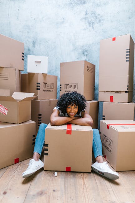 Two individuals involved in packing and home relocation activities indoors, surrounded by stacked cardboard moving boxes of various sizes made from plain brown material with black handles. The man on