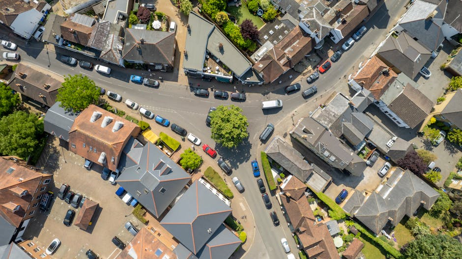 An aerial view of a residential area in Ickenham showing a roundabout at the intersection of several streets, with multiple cars navigating the roadways and parked along the curbs. Surrounding the roa