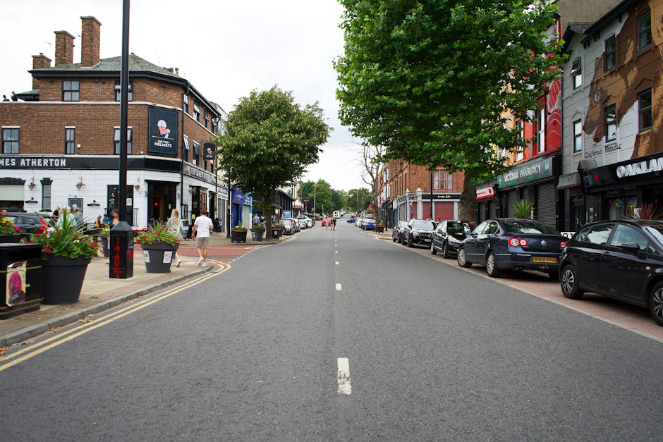 A wide view of a street scene in Ickenham showing a straight asphalt road lined with parked cars on both sides and pedestrian pavement on each sidewalk. On the left side, there is a two-storey brick b