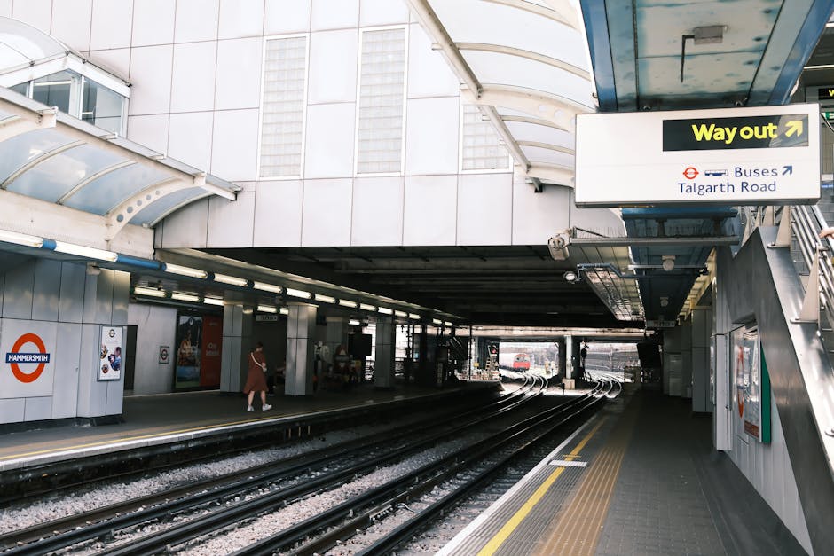 The image depicts the platform area of Hammersmith London underground station during daytime, with the station's signage visible on the left side. The platform has a few passengers, including a woman in a brown coat walking toward the exit, and is equipped with accessibility features like tactile paving along the edge for safety. The station's curved, transparent roof allows natural light to illuminate the space, and the surroundings include tiled walls, overhead lighting, and various informational signs. There are multiple train tracks running through the platform, with some visible trains in the background, indicating active service. The station interface features directional signs, including an overhead sign indicating the way out towards Talgarth Road and bus links, showcasing the station's role as a transportation hub. As part of a house removal or relocation process, [COMPANY_NAME] might use such a station environment for transporting furniture or boxes during a professional moving service, reflecting the logistics involved in home relocations in London.