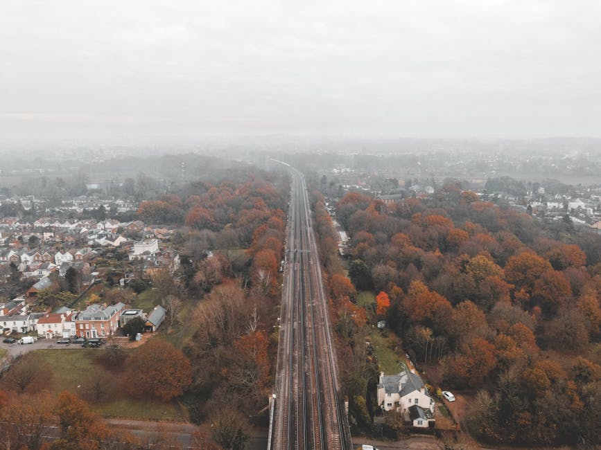An aerial view of a railway line cutting through a suburban area surrounded by trees showing autumn foliage with orange, red, and brown leaves. Residential houses with gardens line both sides of the tracks, which extend into the distance under a cloudy sky. On the left side, there are small houses and driveways, while on the right side, larger homes and wooded areas are visible. The scene captures the quiet environment typical of a home relocation route, with no people or moving equipment in sight, focusing on the landscape and infrastructure relevant to furniture transport and packing during a house move. The image emphasizes the transportation route used during moving services offered by Man With a Van Ickenham, aligning with the context of a moving guide from Ickenham station to Swakeleys Road.