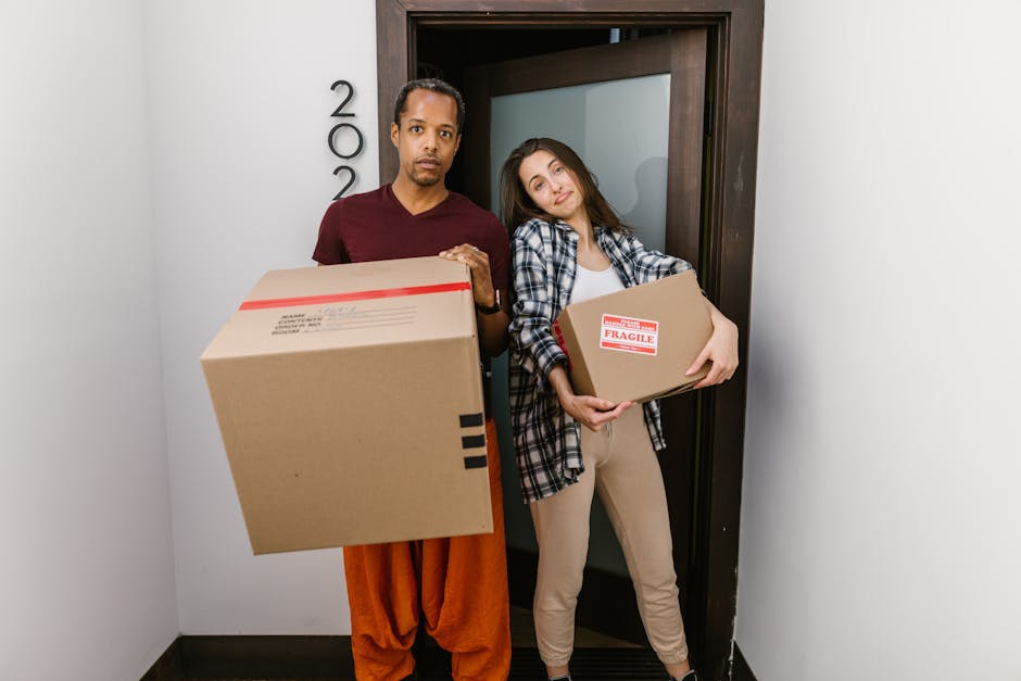 A woman with long, curly hair is sitting inside a room during a house removal, holding a notepad and pen while taking notes. She is wearing a light grey cardigan over a white top. Behind her, a man wi